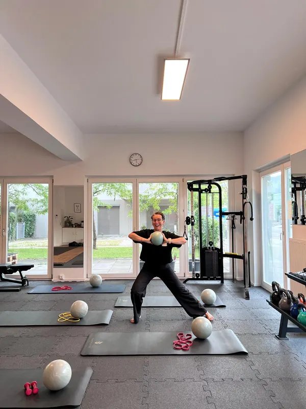 Tini zeigt im Trainingsraum eine Übung mit dem Ball, davor vorbereitete Matten mit Bällen und Bändern