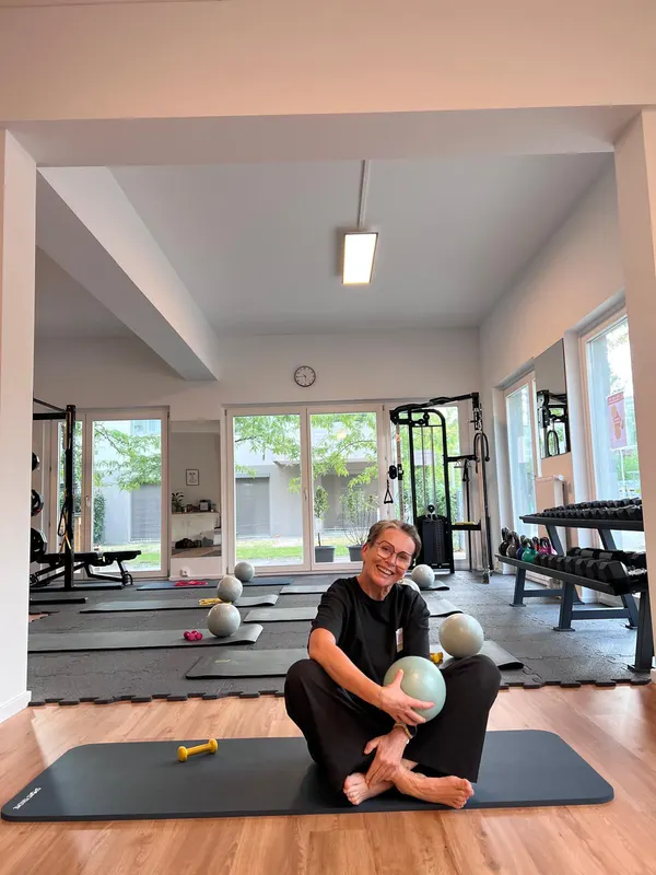 Tini sitzt lächelnd mit einem Ball auf einer Matte im hellen Trainingsraum